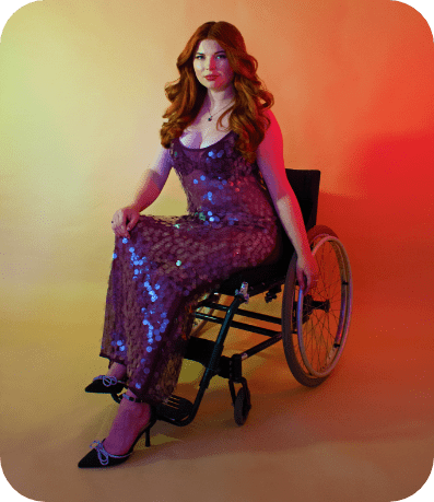 A professional modelling shot of Jenni sat in her wheelchair, wearing a dazzling giant sequin purple iridescent dress. She is sat against a light yellow/orange ombre background.