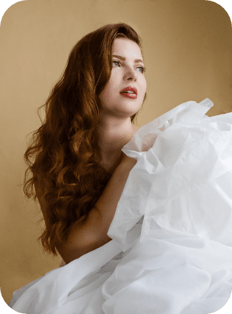 Jenni looking to the right, covered in soft white fabric. Her long auburn hair trails her shoulder with soft curls, complementing her neutral, rosy makeup.