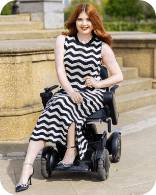 Jenni sitting in her wheelchair, looking towards the camera smiling. She is wearing a black and white chevron dress with black heels.