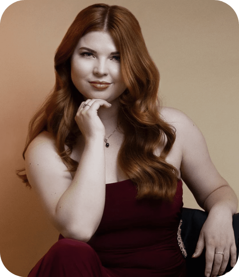 A professional model shot of Jenni looking at the camera and slightly smiling. She has long auburn hair which is curled and is wearing a red dress.